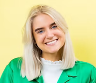 Woman with blonde hair wearing a green jacket and smiling against a yellow background.